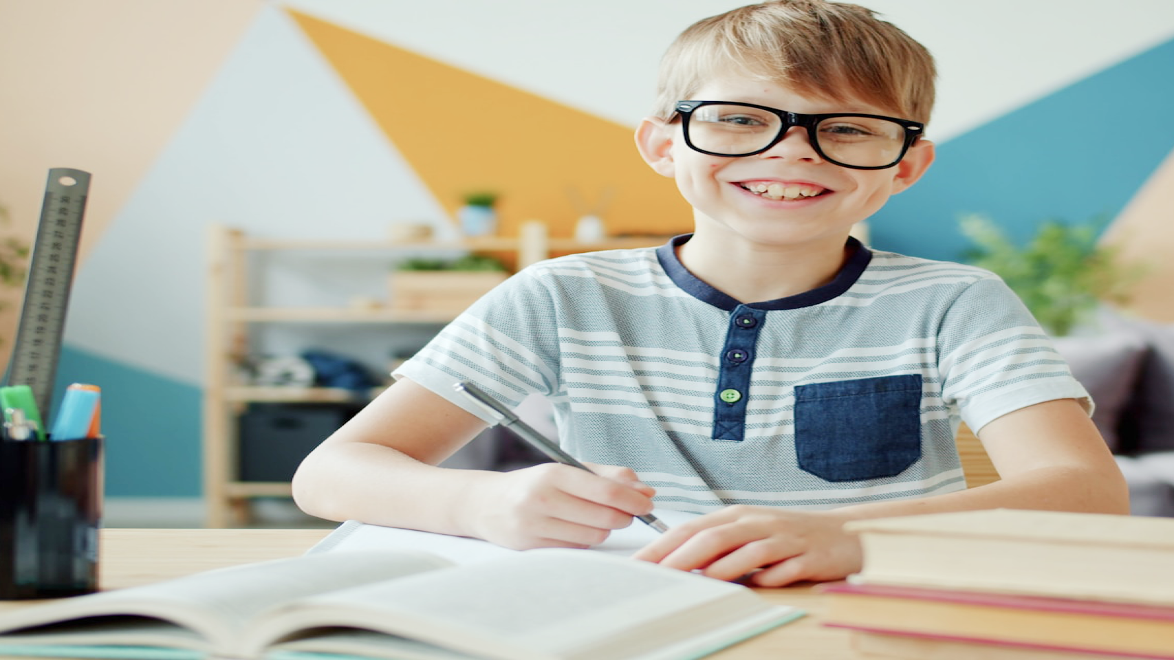 A young boy with glasses smiles while writing in a notebook at a desk, school supplies beside him.